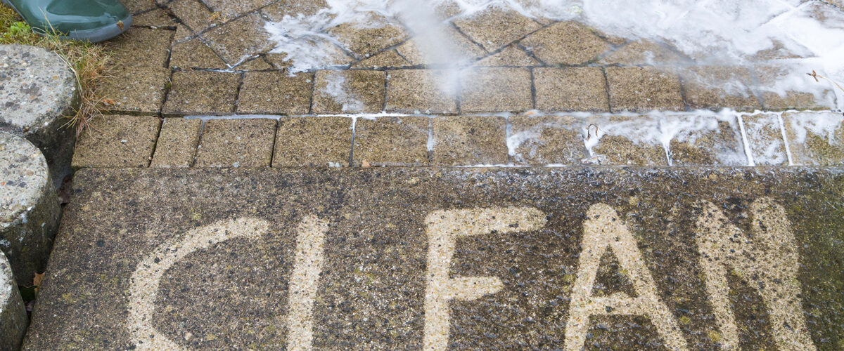 A person uses a pressure washer to clean paving stones, revealing the word "CLEAN" on a dirty surface—demonstrating how pressure washing can refresh your home exterior. A person uses a pressure washer to clean paving stones, revealing the word "CLEAN" on a dirty surface—demonstrating how pressure washing can refresh your home exterior