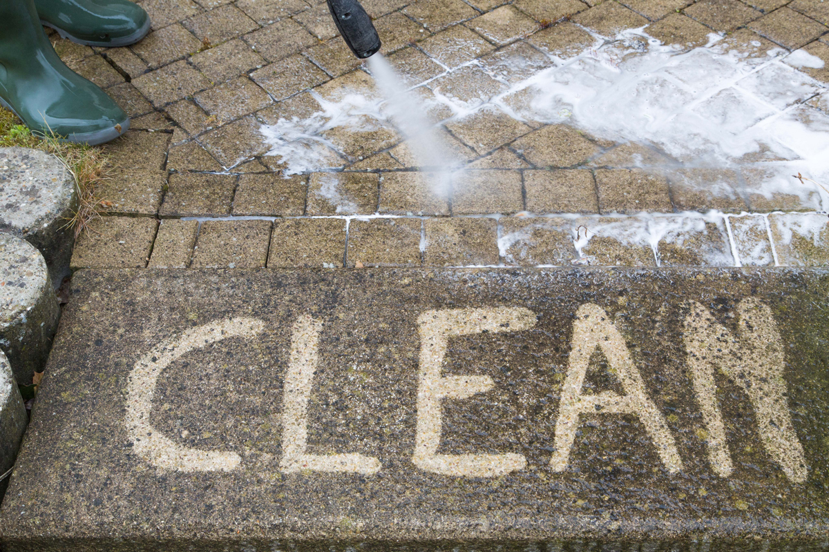 A person uses a pressure washer to clean paving stones, revealing the word "CLEAN" on a dirty surface—demonstrating how pressure washing can refresh your home exterior. A person uses a pressure washer to clean paving stones, revealing the word "CLEAN" on a dirty surface—demonstrating how pressure washing can refresh your home exterior.