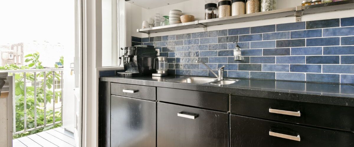 Modern kitchen with black cabinets, blue tiled backsplash, double sink, and various jars on shelves; a door opens to a small balcony with greenery outside. Modern kitchen with black cabinets, blue tiled backsplash, double sink, and various jars on shelves; a door opens to a small balcony with greenery outside