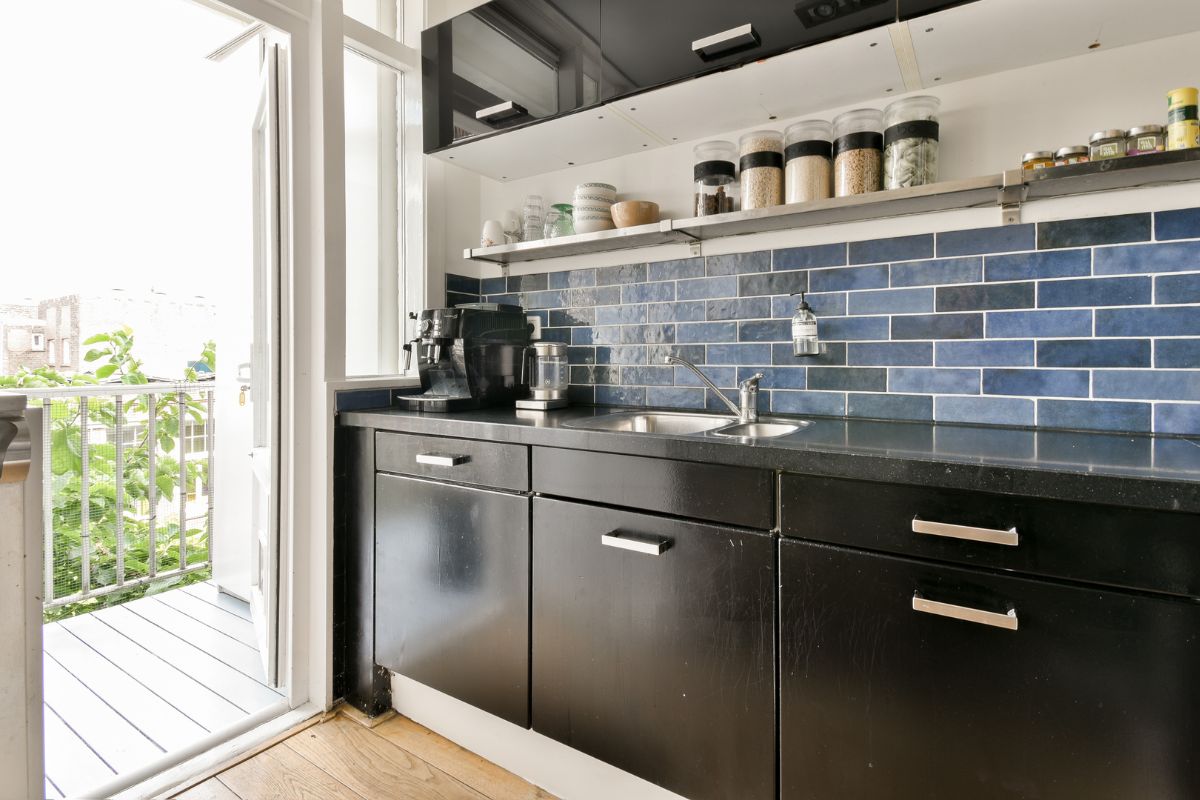 Modern kitchen with black cabinets, blue tiled backsplash, double sink, and various jars on shelves; a door opens to a small balcony with greenery outside. Modern kitchen with black cabinets, blue tiled backsplash, double sink, and various jars on shelves; a door opens to a small balcony with greenery outside